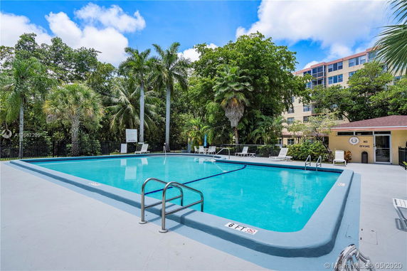 Outdoor swimming pool area with lounge chairs and surrounded by trees, adjacent to a multi-story building.