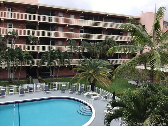 View of a multi-story building with balconies, surrounded by palm trees, and a swimming pool in the foreground.