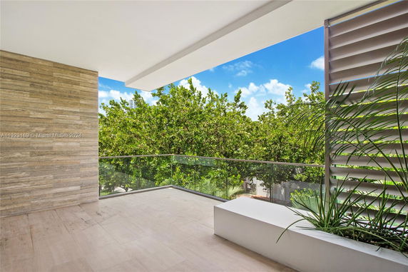Outdoor balcony with glass railing overlooking green trees, featuring a modern design with a beige tiled floor and partial wooden wall.