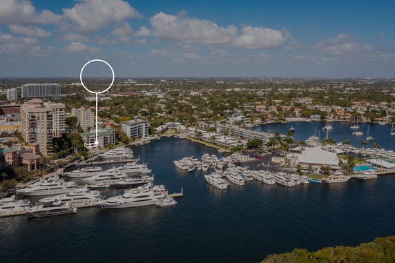 View of a marina with multiple yachts docked and a surrounding suburban area with residential and commercial buildings.