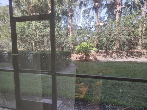 View of a garden area with grass, bushes, and tall trees seen from inside a building through glass doors.