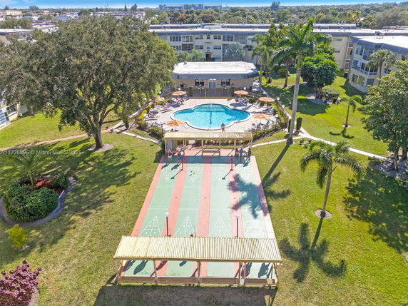 Aerial view of a swimming pool area with a surrounding deck and adjacent shuffleboard court in a residential garden.