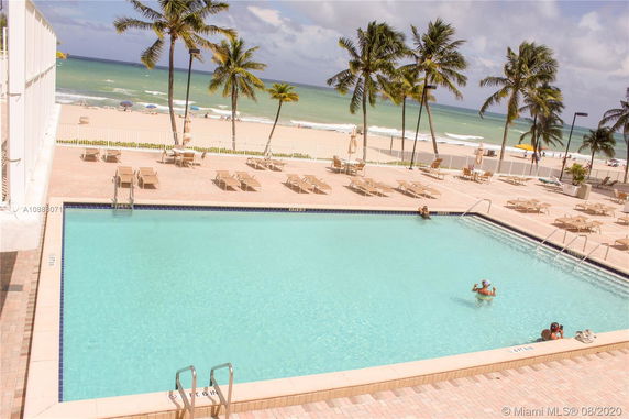 Outdoor swimming pool area with sun loungers, overlooking a beach with palm trees and ocean.