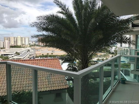 Balcony view with a glass railing, overlooking a cityscape with a large palm tree and distant buildings.