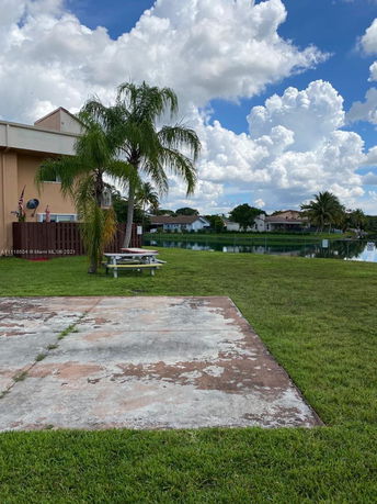 Side view of a building near a grassy area and a pond, with palm trees and cloudy skies.