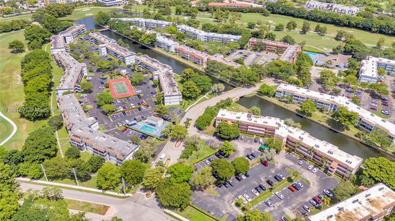 Aerial view of a residential area with multiple apartment buildings, surrounded by greenery and water bodies, including parking lots and a tennis court.