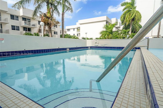 Outdoor swimming pool area with palm trees and modern buildings in the background.