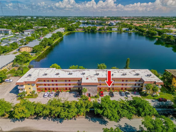 Aerial view of a multi-story building near a lake with a parking area in front.