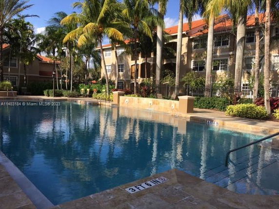 Outdoor swimming pool with surrounding palm trees and a multi-story building in the background.