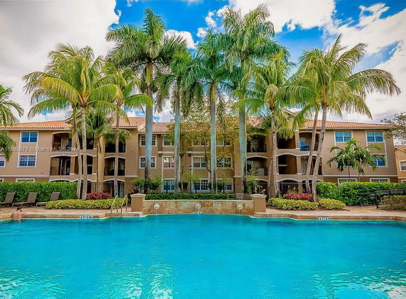 Outdoor swimming pool area with a three-story residential building and palm trees in the background.