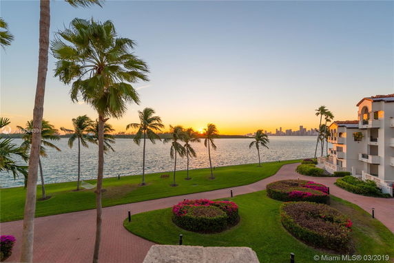 View of a waterfront with palm trees and a distant city skyline at sunset.