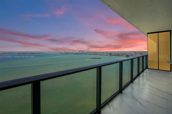 Balcony with glass railing offering a panoramic view of the ocean and city skyline at sunset.