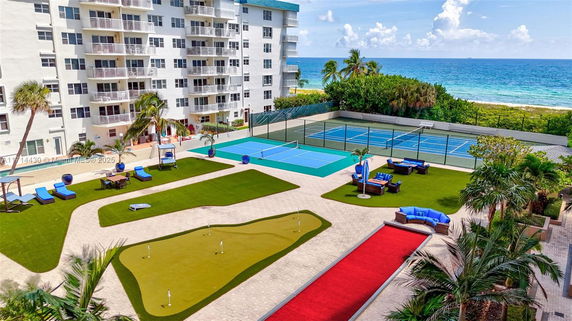 Outdoor area with a tennis court and lounging spaces near a multi-story building by the ocean.