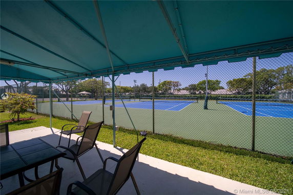 Tennis court viewed from a shaded seating area with chairs and tables.