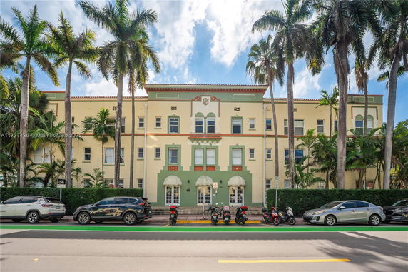 Front view of a three-story building with palm trees and parked vehicles.