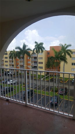 View from a balcony overlooking multi-story residential buildings with palm trees and a parking area.
