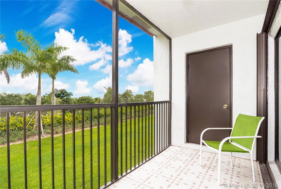 Enclosed balcony with metal railing and green chair, overlooking grass and palm trees.
