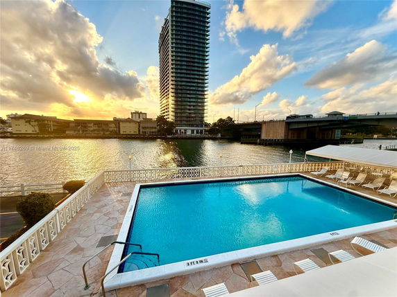 Outdoor swimming pool with surrounding lounge chairs near waterfront and high-rise building in the background.