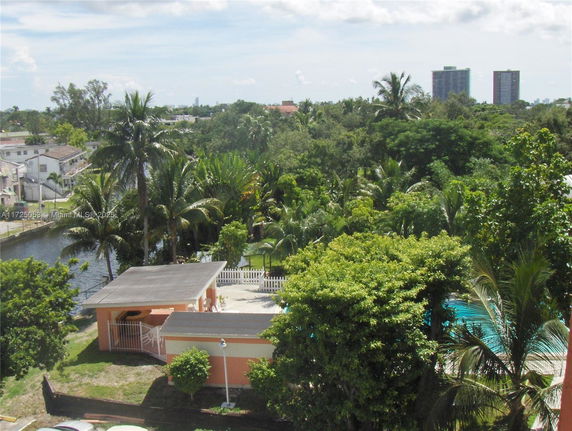 Panoramic view of a lush green area with tall palm trees, a small building, and distant high-rise buildings.