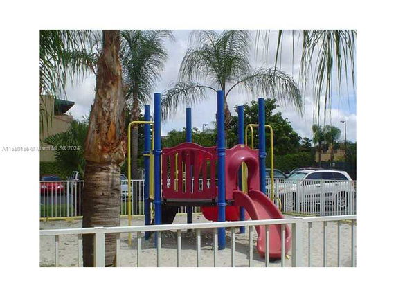 Outdoor playground with a slide and climbing structure, surrounded by palm trees and a fence.