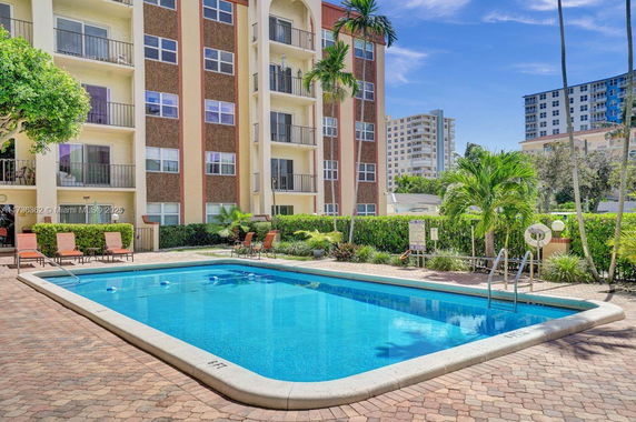 Outdoor swimming pool area with surrounding buildings and palm trees.