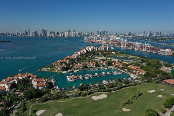 Aerial view of a coastal area with residential buildings, a marina with yachts, and a golf course. The skyline of a city and a busy port are visible in the background across the water.