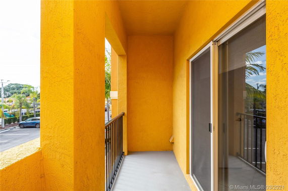 View from a yellow-painted balcony with black railings, overlooking a parking area and street.