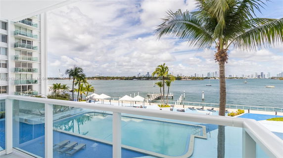 Panoramic view of a waterfront area with a swimming pool, palm trees, and distant city skyline.