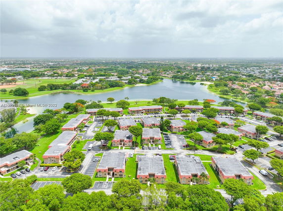 Aerial view of a residential area with multiple low-rise buildings and parking lots, surrounded by greenery and a water body.