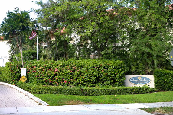 Front view of a condo building partially visible behind trees and bushes, with a sign reading 'Ocean Village Condominiums'.