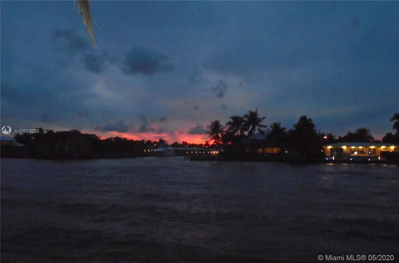 View of a waterfront area at sunset with silhouetted palm trees and lights from a building.
