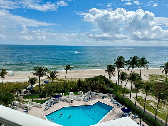 View of a swimming pool near a beach with palm trees and ocean in the background.