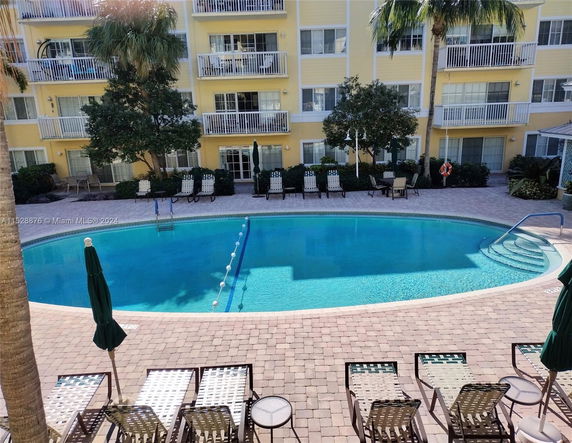 View of an outdoor swimming pool with surrounding chairs and a yellow multi-story building in the background.