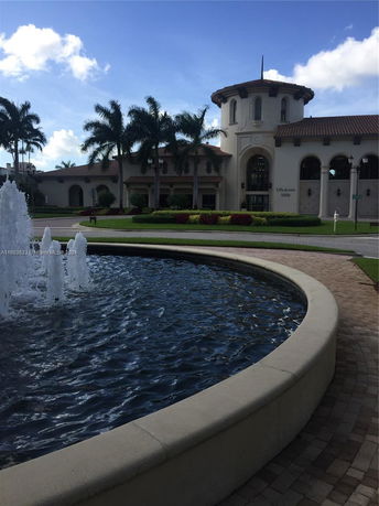 Front view of a large building with arched windows and a cylindrical tower, surrounded by palm trees and a fountain.