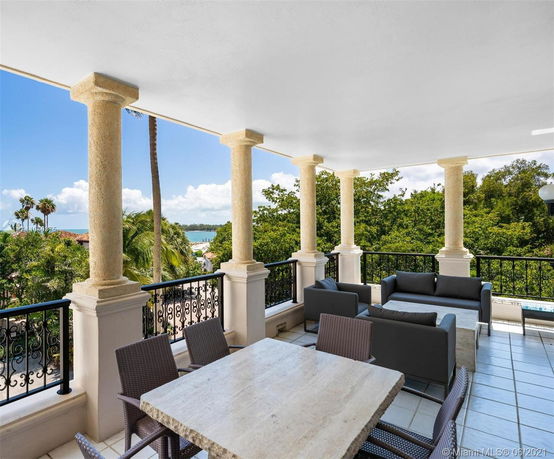 Covered outdoor seating area with stone columns and patio furniture, offering views of lush greenery and distant water.