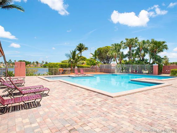 Outdoor view of a swimming pool area with a paved deck and lounge chairs, surrounded by greenery and a clear blue sky.