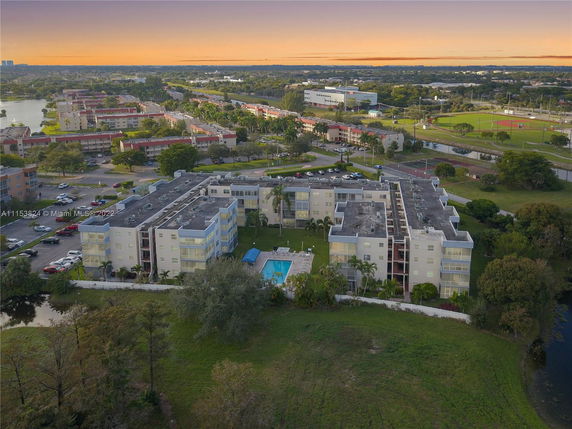 Aerial view of a large residential building complex with a swimming pool, surrounded by greenery and other residential buildings, under a colorful sunset sky.