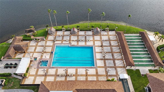 Aerial view of an outdoor swimming pool complex with a large rectangular pool surrounded by lounge chairs and palm trees, adjacent to a body of water.