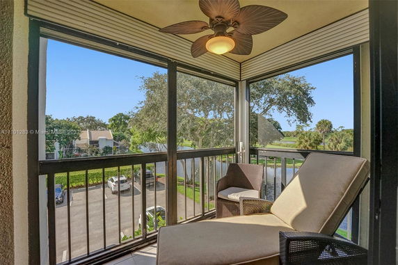 Outdoor patio area with a relaxing chair and fan, overlooking a calm grassy and treed landscape with nearby parking.