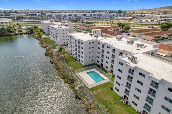 Aerial view of a large multi-story apartment complex next to a body of water, with a swimming pool visible within a fenced area.