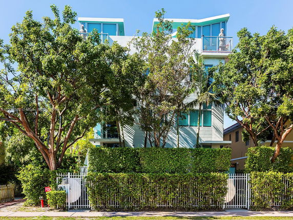 Front view of a multi-story modern building with balconies and large windows, surrounded by trees and a fence.