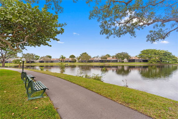 View of a pond with benches and a path running alongside it, trees and houses in the background.