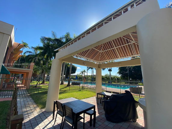 Outdoor area with a covered seating space and a view of a swimming pool surrounded by a fence.