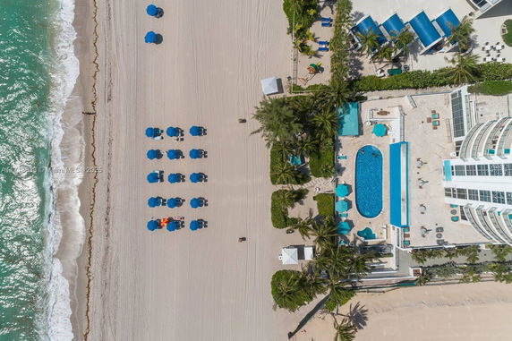 Aerial view of a beachfront property with a swimming pool and beach setup with umbrellas and loungers.