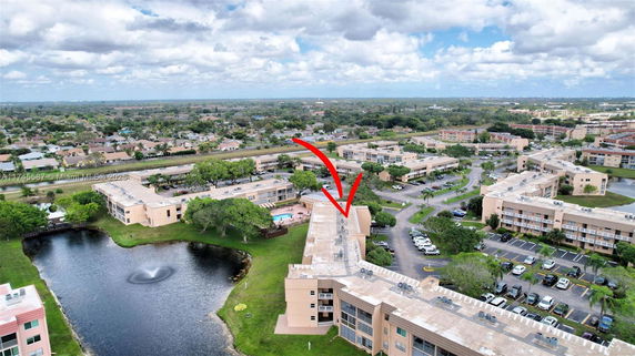 Aerial panoramic view of a residential area with multiple apartment buildings and a parking area, surrounded by greenery and a pond.