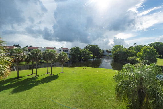 Scenic view of green landscape with palm trees and a pond, buildings in the distance under a cloudy sky.