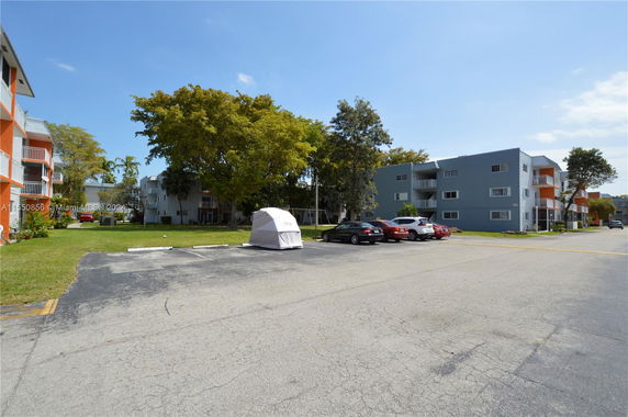 Outdoor parking area with several cars and multi-story buildings in the background.
