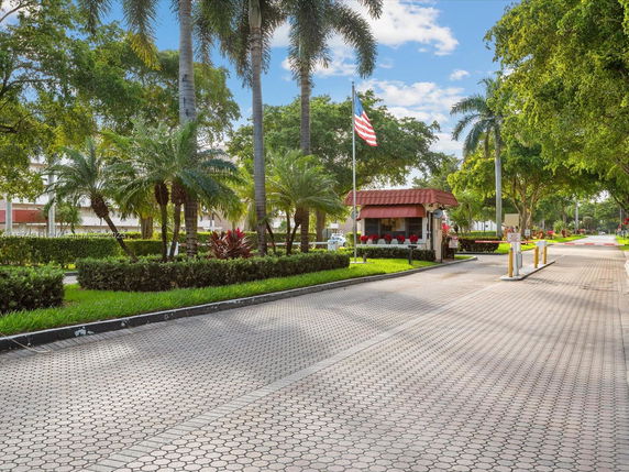 Entrance area with a flagpole and small security booth surrounded by palm trees.