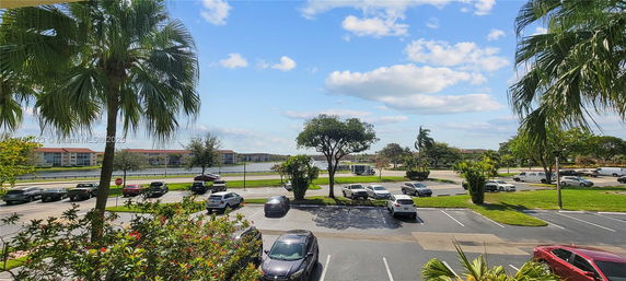 View of a parking area with several cars, palm trees, and buildings in the distance under a blue sky.