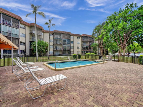 Outdoor swimming pool area with lounge chairs and a multi-story building in the background.